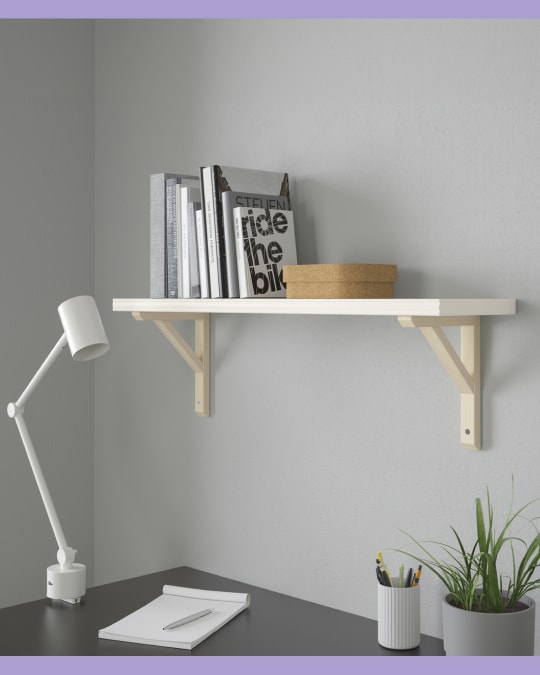 Repisa flotante blanca con soportes de madera clara sobre un escritorio; contiene libros, dos cajas redondas y un objeto decorativo.