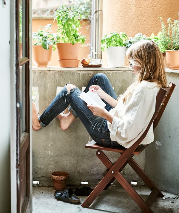Rhianna นั่งอยู่ที่เก้าอี้ไม้พับได้และอ่านหนังสือที่ระเบียงด้านนอกซึ่งมีกระถางต้นไม้ตั้งอยู่