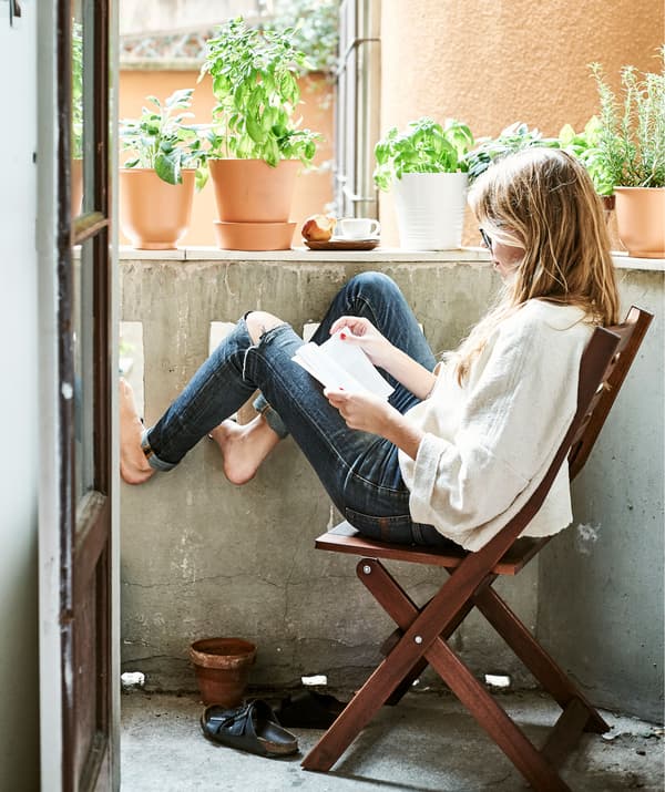 Rhianna sitting on a foldaway wooden chair and reading on her outside balcony lined with plants in pots.