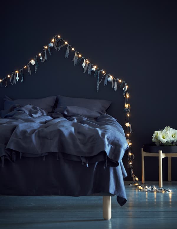 Ribbons and baubles tied to a light chain and pinned on a wall, above a bed.