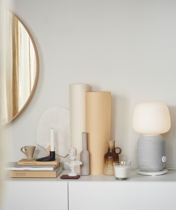 Ripiano di un mobile con libri, una candela profumata, una lampada con cassa Wi-Fi e oggetti decorativi in ceramica. A lato, uno specchio alla parete - IKEA