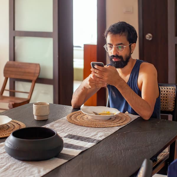 رجل ذو شعر داكن ولحية انتهى للتو من تناول الطعام ويقوم بتصفح هاتفه المحمول على طاولة المطبخ.