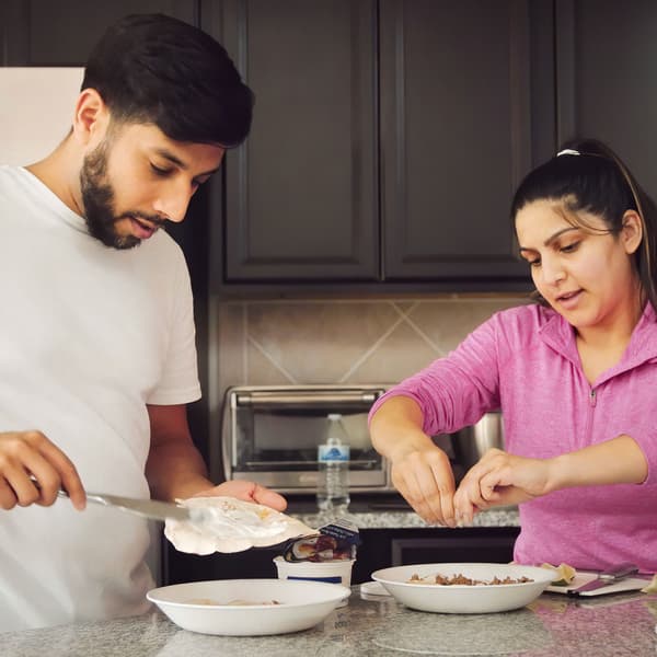 رجل وامرأة أمام كل منهما طيق، يقومان بتحضير الإفطار في مطبخ به سطح عمل حجري وخزائن رمادية.