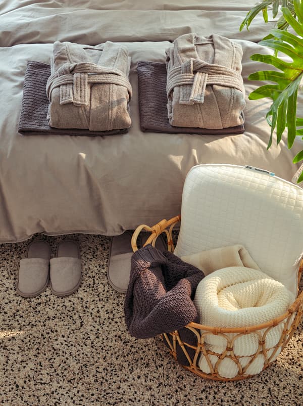 เสื้อคลุมอาบน้ำ ROCKÅS พับไว้อย่างเรียบร้อยแบบโรงแรม และผ้าเช็ดตัวใหม่วางอยู่บนเตียง ตะกร้า SNIDAD/สนีดัด ใส่ผ้าคลุมและหมอน