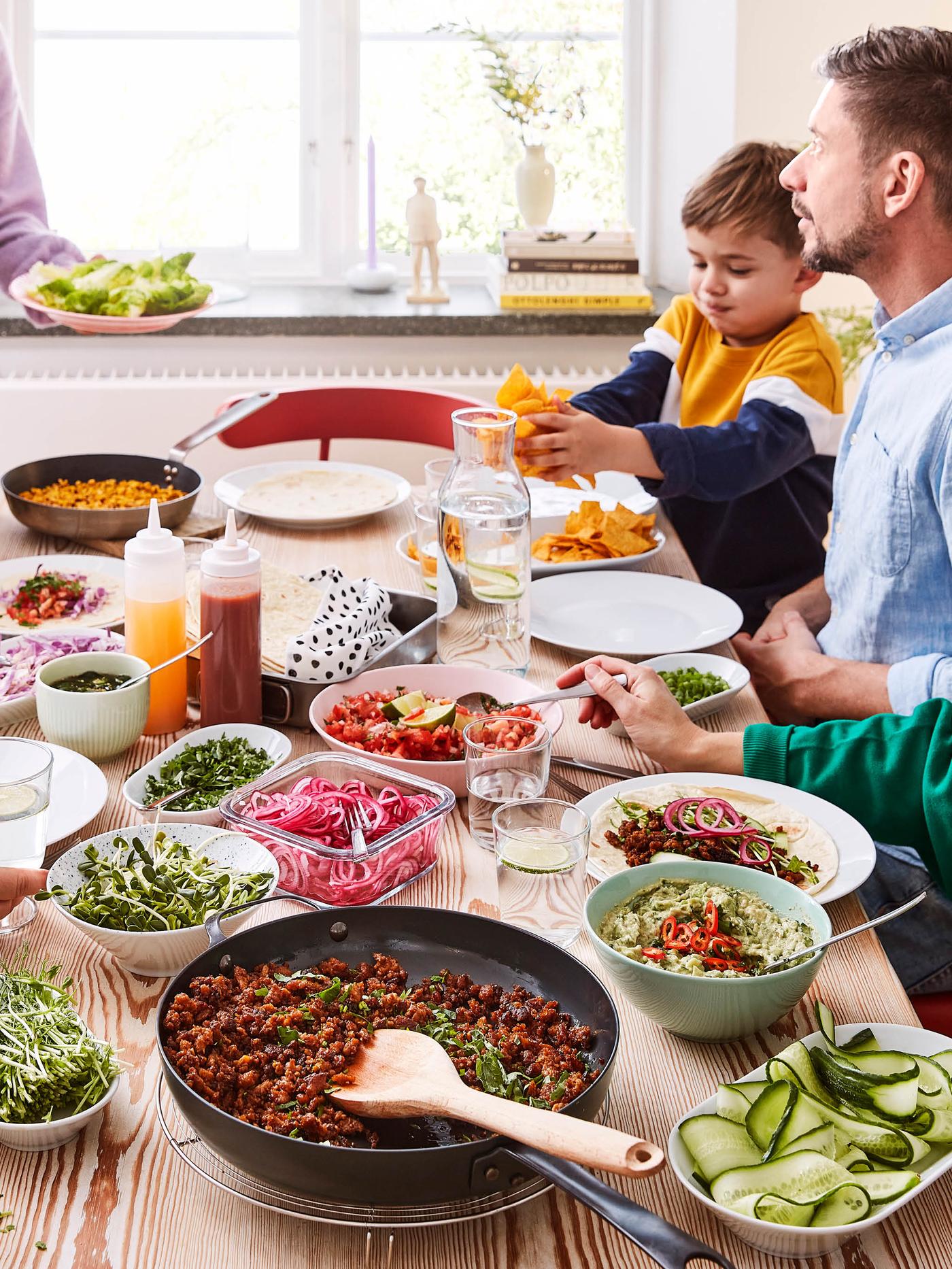 Rodina sedí pri stole plnom tanierov a misiek s jedlom, vpredu je panvica VARDAGEN s bezmäsitou zmesou VÄRLDSKLOK na tacos.