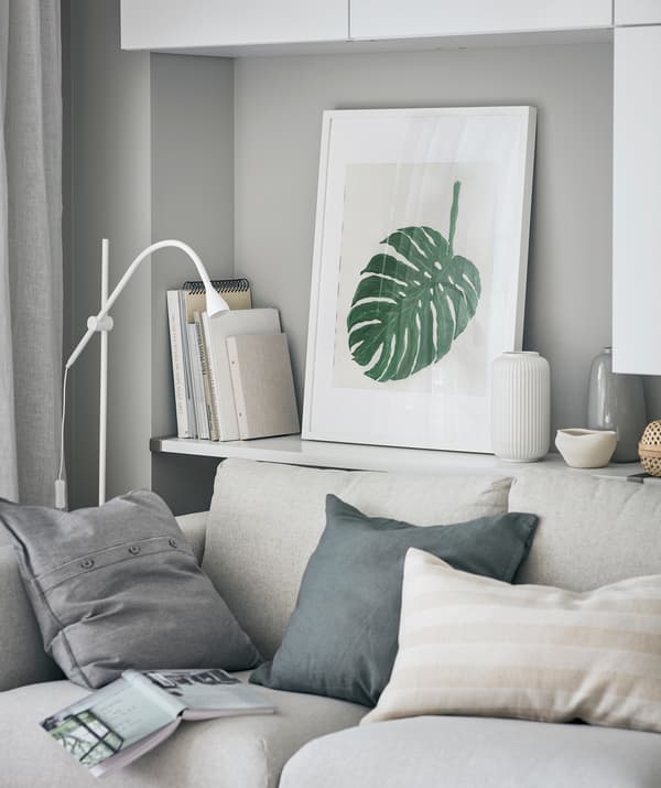 Roh obývacej izby s nástennou policou za pohovkou s knihami, dekoráciami a zarámovanou kresbou listu rastliny monstera. 