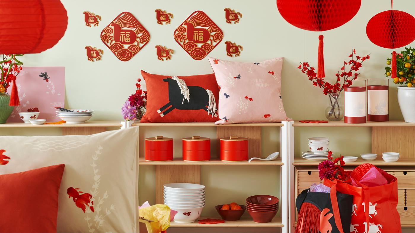 Room decorated for Lunar New Year with red lanterns, cushions, festive wall hangings, dishes, and gift bags arranged on wooden shelves and tables.