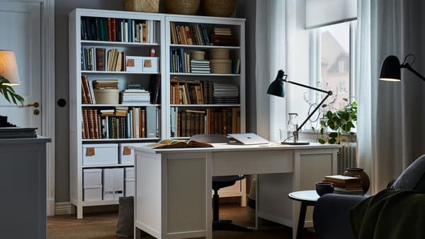 Room with a white desk placed centrally, matching tall bookcases behind the desk and a work lamp on the desk.