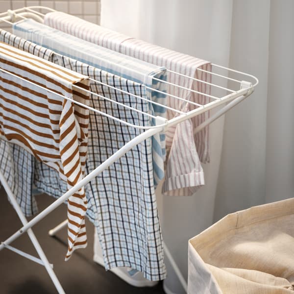 Ropa a rayas y cuadros colgada en un tendedero JÄLL blanco frente a una cortina de baño blanca, detrás de una bolsa de lavandería beige.