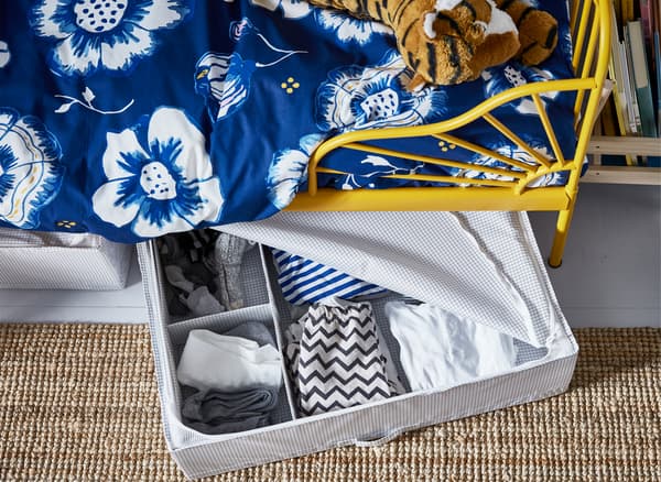 Ropa en una caja de almacenaje abierta debajo de una cama infantil con estructura amarilla y ropa de cama con motivos florales azul.