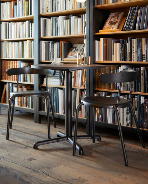 Round anthracite café table with hooks for bags underneath, two chairs, in front of a large bookcase filled with books.