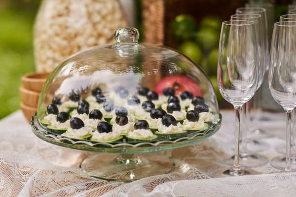 Round glass bowl with lid full of snacks next to glass champagne glasses.