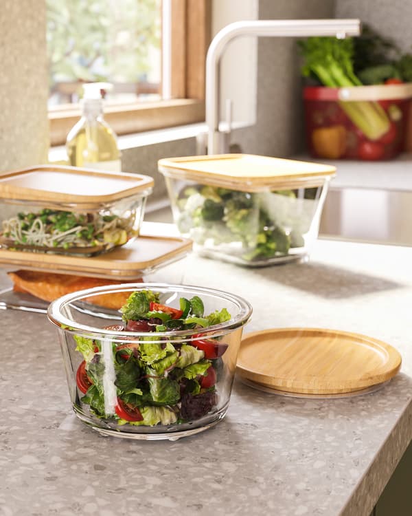 Round glass food containers with tight-fitting bamboo lids with vegetables inside, they're standing on a kitchen countertop.