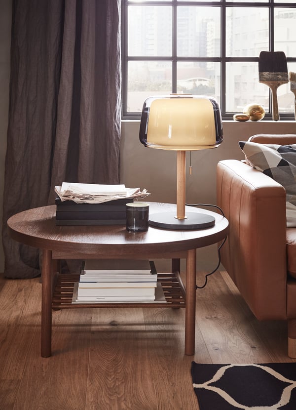 Round wooden LISTERBY coffee table with a lamp and papers by the sofa.