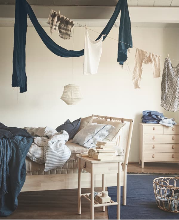 Roupa colgando sobre unha cama con estrutura de madeira, unha mesa de noite e unha cómoda con caixóns.