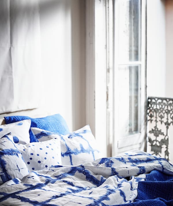 Roupa de cama TÄNKVÄRD con motivos azuis colocada en capas sobre unha cama ao lado da porta dun balcón.