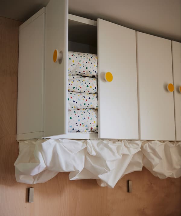 Row of closable wall cupboards, top reaching the ceiling. Stacked duvets within, cloud-like billows of fabric underneath.