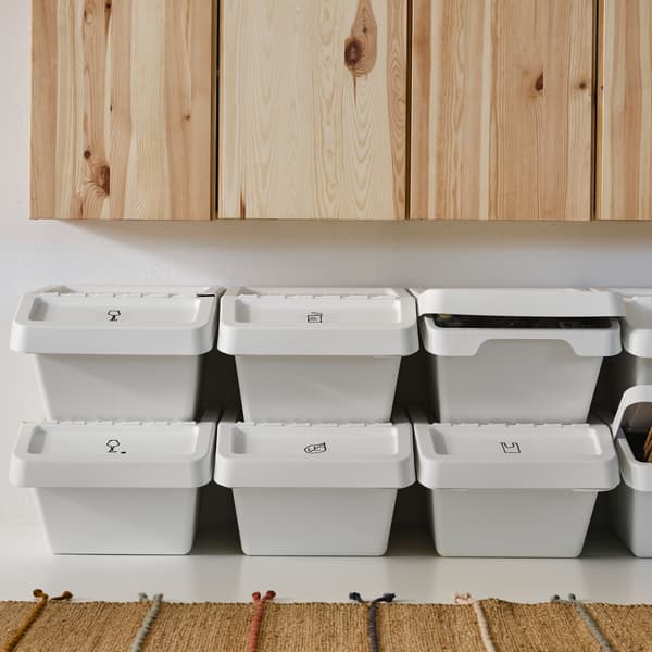 Rows of white SORTERA waste sorting bins with lids stacked on top of one another under a row of pine cabinets, plus a rug.