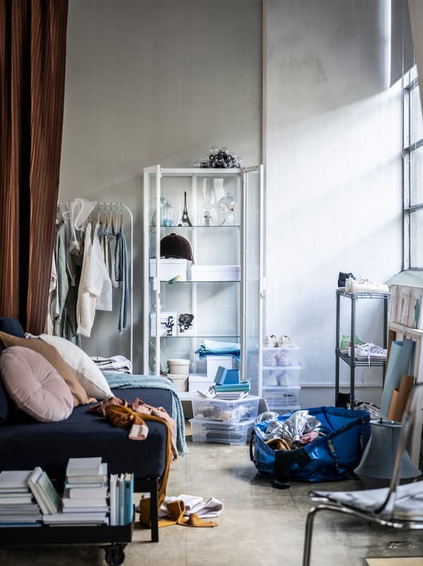 Rozhádzaná izba s pohovkou, bielym policovým dielom, závesom, knihami a modrou taškou IKEA na podlahe.