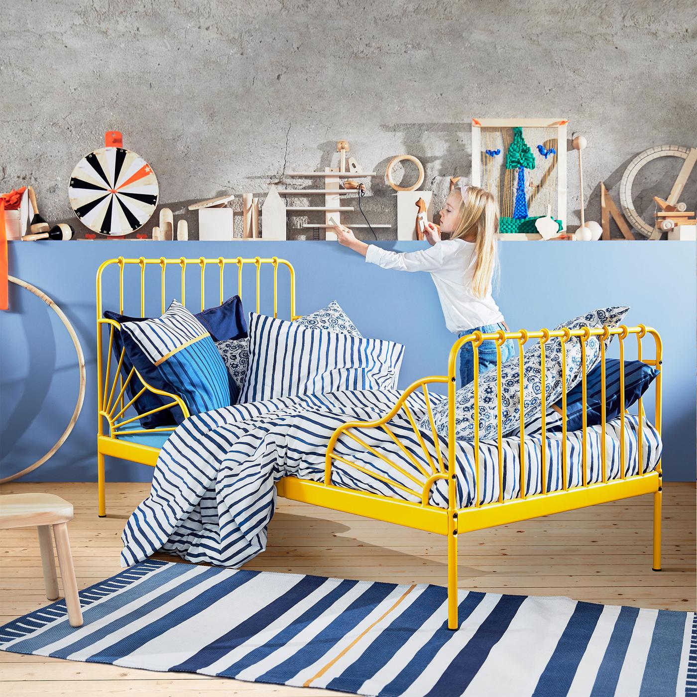 Rozkládací postel MINNEN, kterou lze roztáhnout až na 207 cm. Je práškově nalakovaná na tmavě žlutou barvu a opatřená detaily, které připomínají sluneční paprsky.