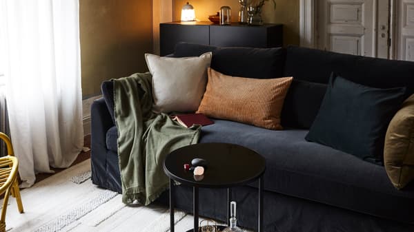 Ruang tamu dengan sofa, kusyen, alas, ambal, meja kecil, saidbod dengan lampu, vas dan kerusi berlengan rotan.