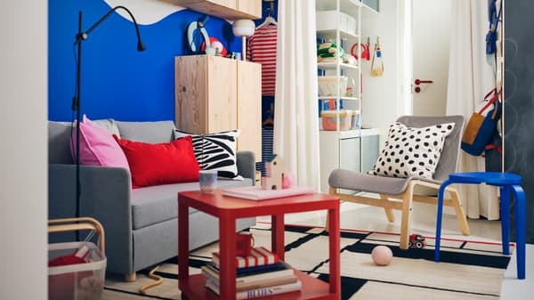 Ruang tamu kecil yang berwarna-warni yang mempunyai dinding berwarna biru terang, katil sofa FIRDHULT berwarna kelabu muda dan meja sisi beroda TINGBY berwarna merah.
