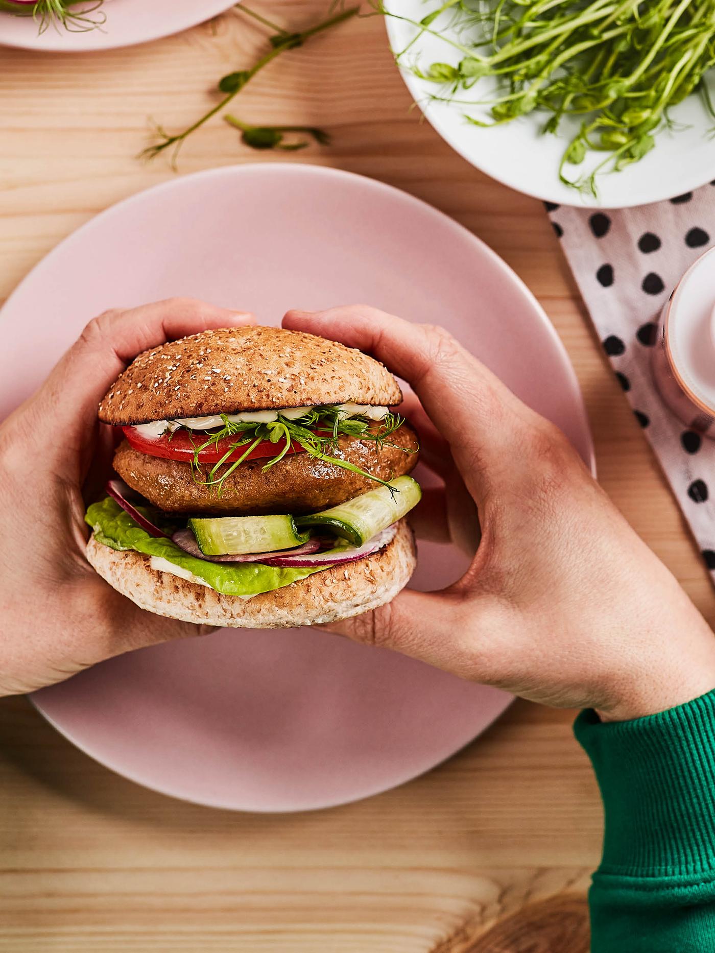 Ruce drží hamburger z bezmasé směsi VÄRLDSKLOK nad růžovým dezertním talířem.