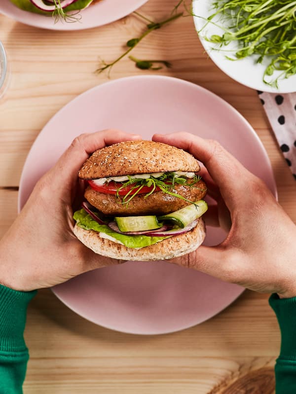 Ruce drží hamburger z bezmasého mletého VÄRLDSKLOK nad růžovým talířem.