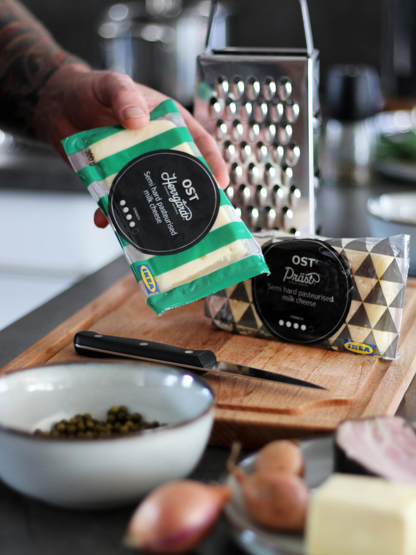 Ruka drží balenie syra OST HERRGÅRD, vzadu je na doske PROPPMÄTT balenie syra OST PRÄST a strúhadlo IDEALISK.