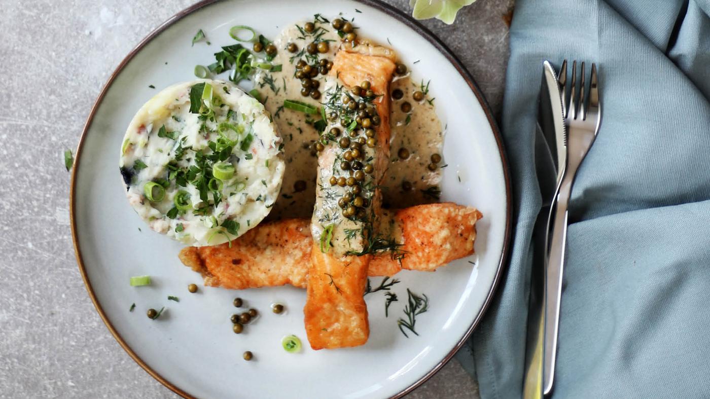 Ruka drží plátek tenkého chleba GRÄDDAT s lososem a salátem, v pozadí na kamenné zídce leží celé balení marinovaného lososa SJÖRAPPORT.