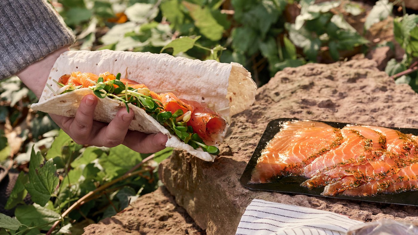 Ruka drží tenkú chlebovú placku GRÄDDAT s lososom a šalátom a v pozadí na kamennom múriku je celé balenie marinovaného lososa SJÖRAPPORT.