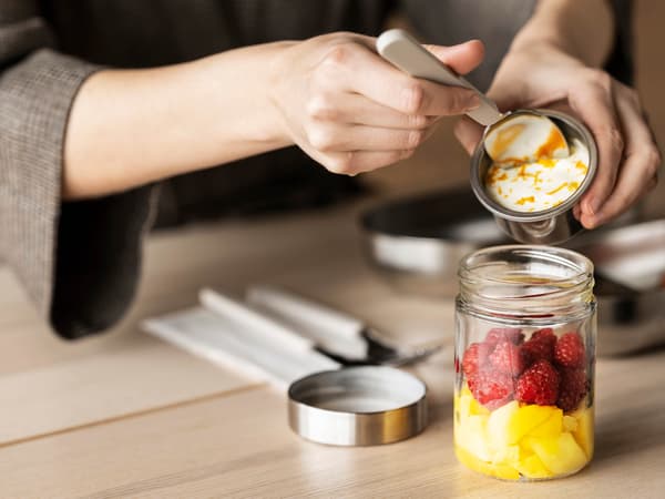 Ruka držící lžíci přidává do dózy DAGKLAR na ovoce jogurt z menší kovové nádobky.