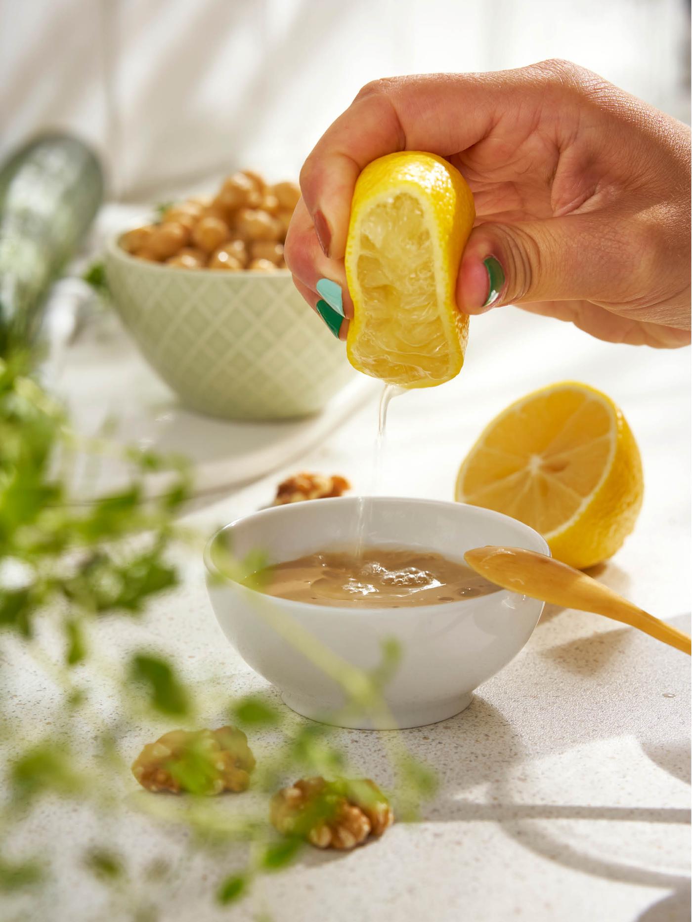 Ruka mačká šťávu z půlky citrónu do misky sezamové pasty tahini, vedle leží druhá půlka citrónu, pár vlašských ořechů a miska cizrny.