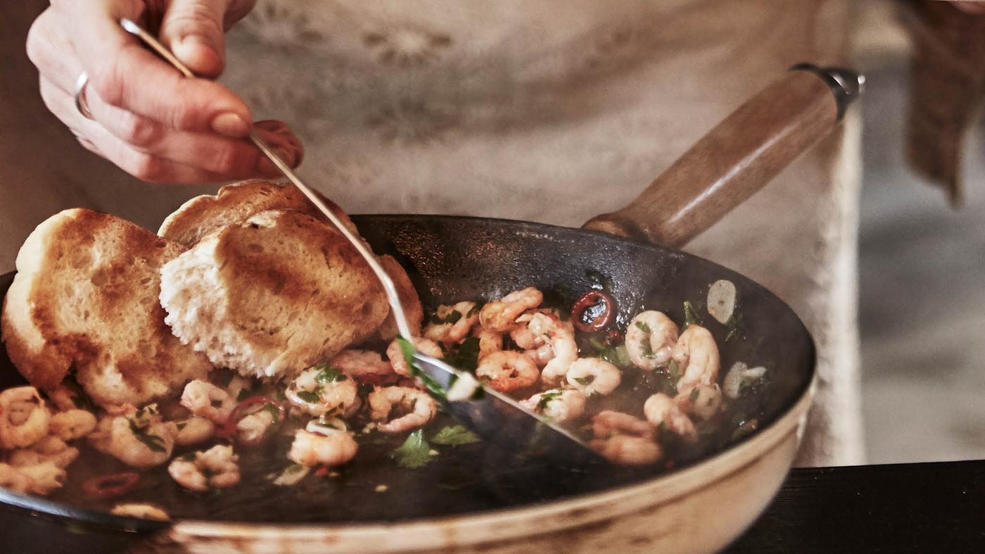 Ruka mieša obracačkou krevety SJÖRAPPORT s petržlenovou vňaťou v liatinovej panvici.