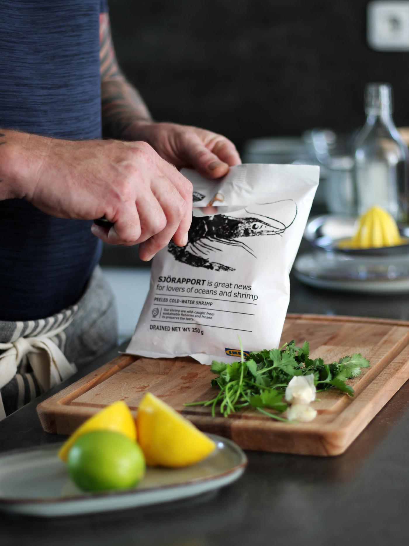 Ruka s nožem načíná sáček krevet SJÖRAPPORT na prkénku PROPPMÄTT  vedle svazečku koriandru, v popředí leží talíř GLADELIG s limetkou a citronem.