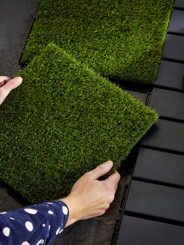 Ruky držia podlahovú krytinu, ktorá pripomína trávnik, nad drevenou podlahou zz tmavohnedého dreva.