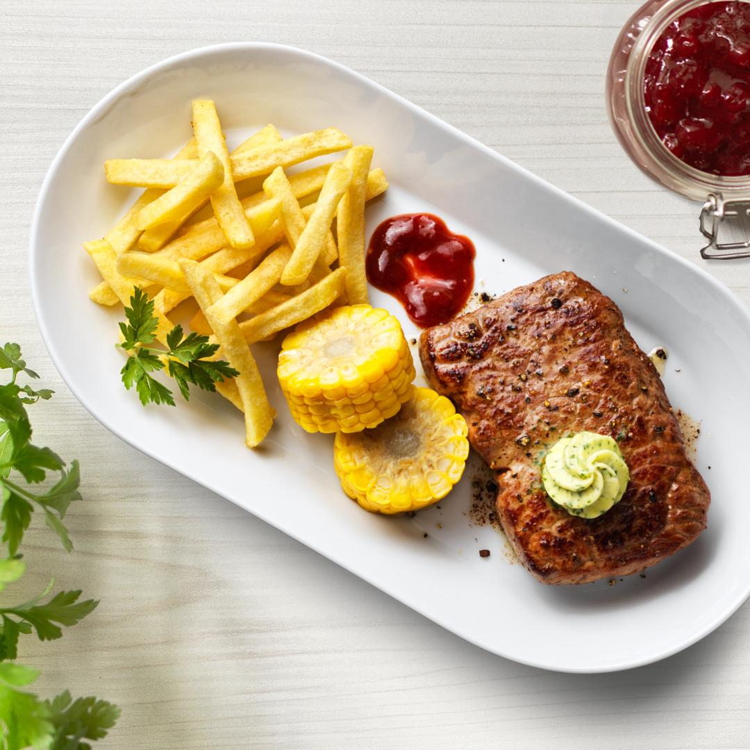 Rumpsteak mit Maiskolben, Pommes und Kräuterbutter auf einem weißen Teller präsentiert.