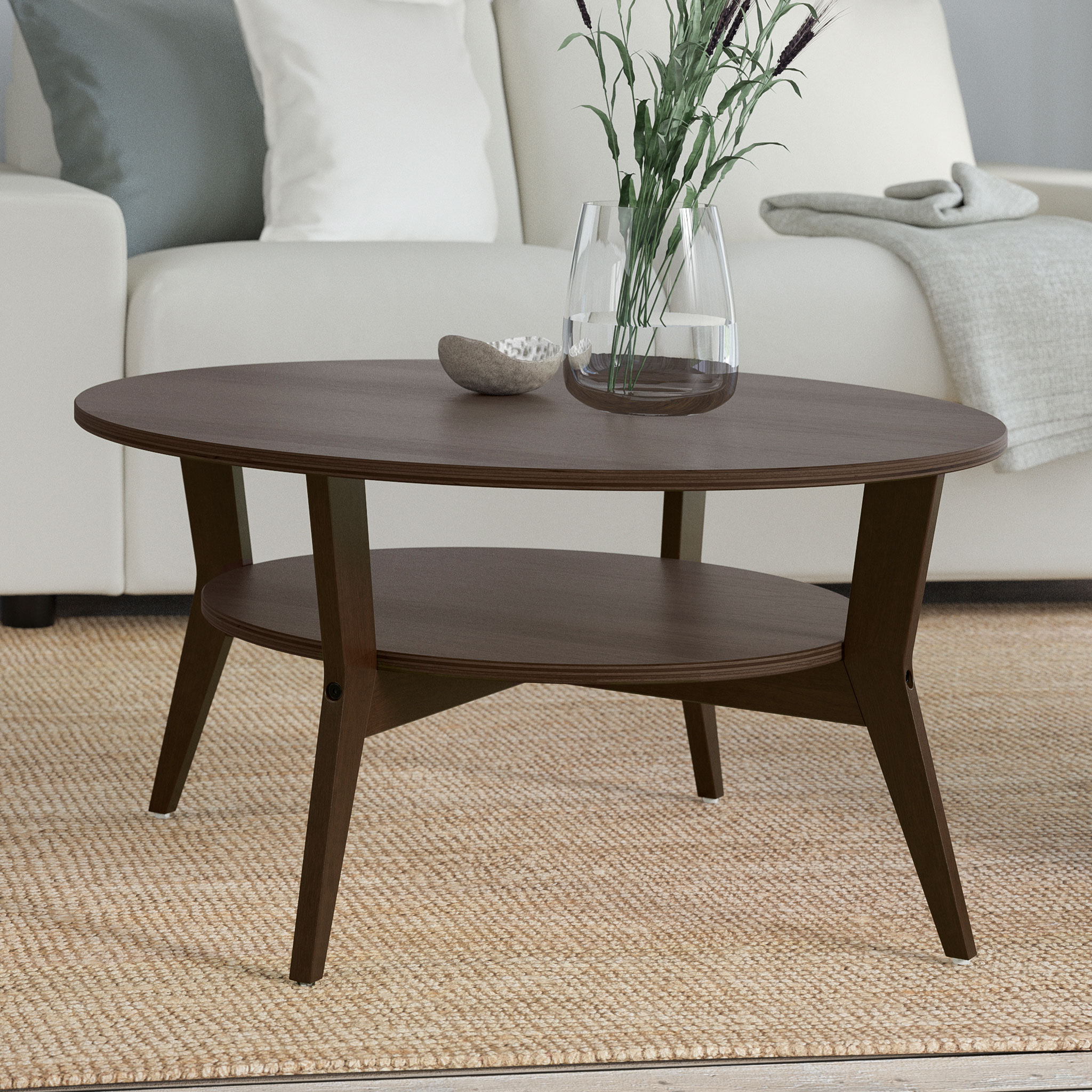 Runt soffbord i mörkbrunt trä med hylla under bordsskivan. Bordet står på en ljus matta framför en soffa, dekorerat med en glasvas med gröna kvistar.