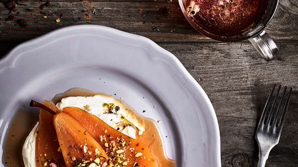 Rustic, wooden table top with a serving of pear-and-dairy-based dessert on a plate; a fork and a cup of tea on the side.
