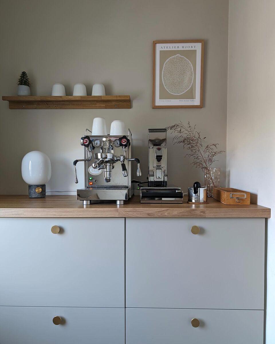 Rustige, minimalistische koffiecorner met een espressomachine en koffiemolen op een lichtgrijs dressoir met houten blad, witte kopjes op een wandplank, een kleine tafellamp, droogbloemen en een ingelijste poster tegen een neutrale muur.