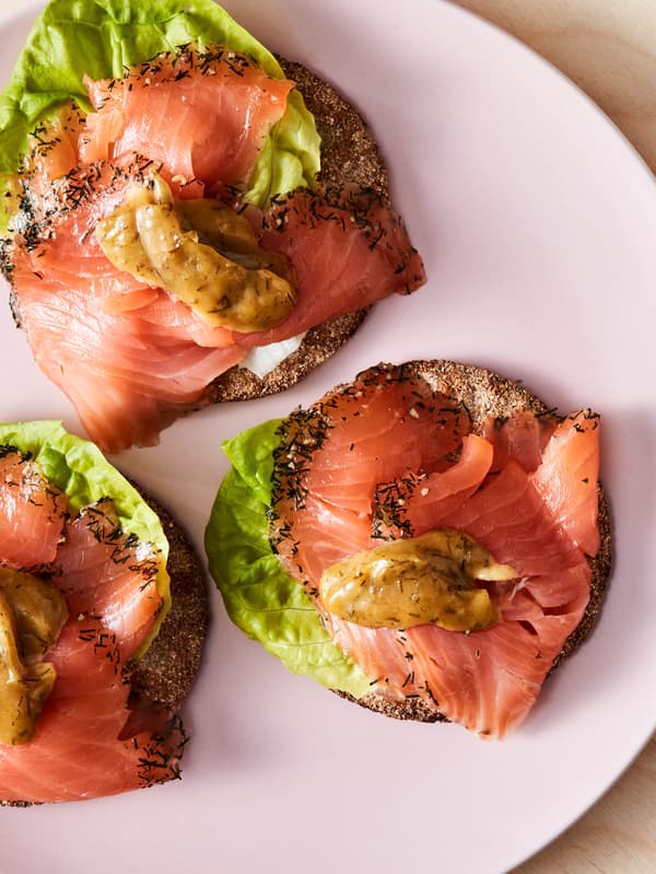 Ružový tanier s tromi kolieskami krehkého chleba KNÄCKEBRÖD FLERKORN ozdobenými listovým šalátom, údeným lososom SJÖRAPPORT a omáčkou na lososa SÅS SENAP & DILL.