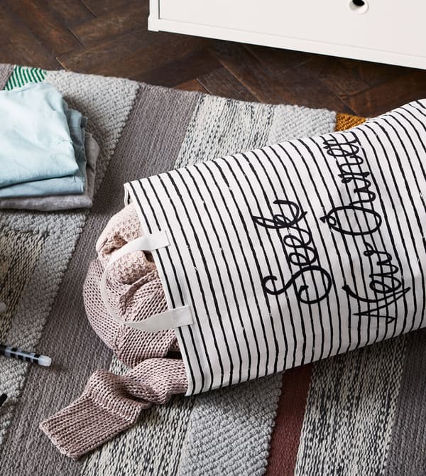 Sacco per il bucato KLUNKA pieno di magliette e una pila di vestiti sparsi su un tappeto grigio - IKEA