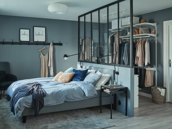 SÄBÖVIK bed in a bedroom draped with blue beddings, resting against a room divider. A closet hanging at the back of the room.