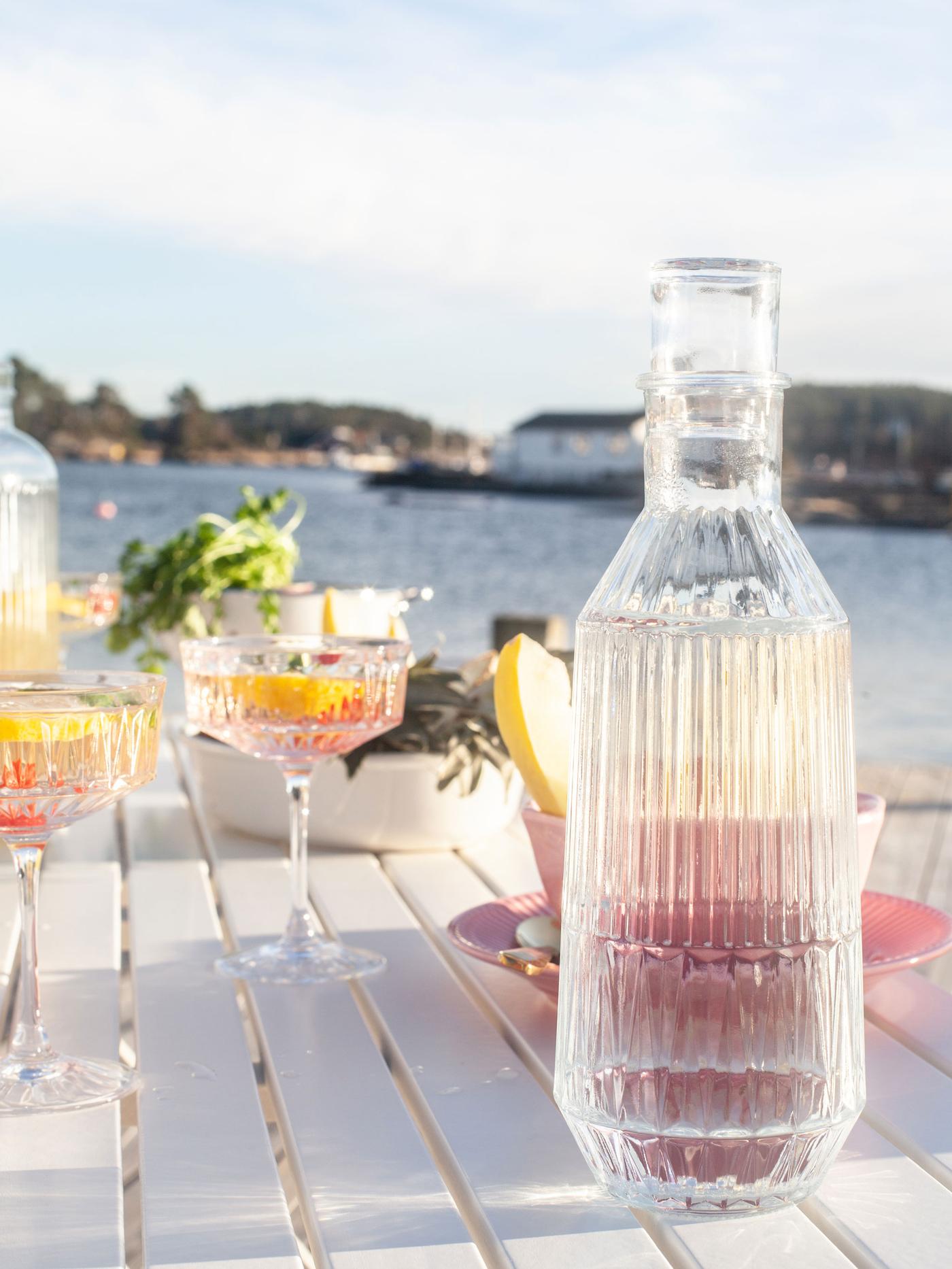 SÄLLSKAPLIG champagnelas og en SÄLLSKAPLIG karaffel står på et hvidt bord med havet i baggrunden.