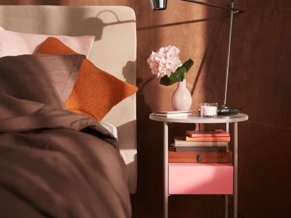 .Säng bredvid ett nattduksbord med böcker och en lampa och en blomma och ett ljus.