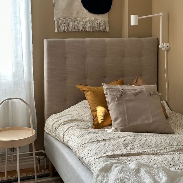 Säng med beige sänggavel, flera kuddar i senapsgult och beige, sidobord, vägglampa och dekorativ väggbonad ovanför.