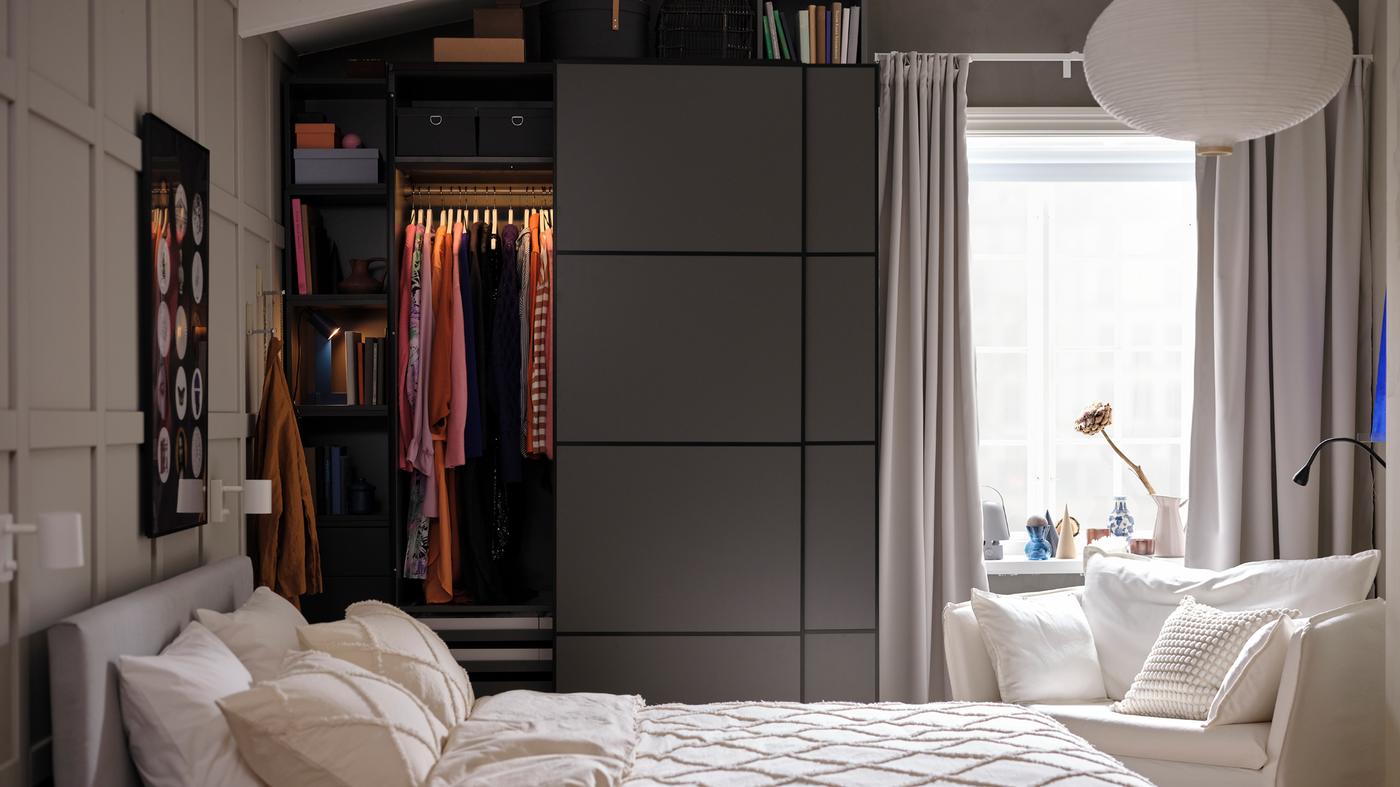 Sängyn takana on tummanharmaa PAX/MEHAMN-vaatekaappi, jonka vasen liukuovi on hieman avoinna. Avoimesta ovesta näkyy henkareille ripustettuja vaatteita.