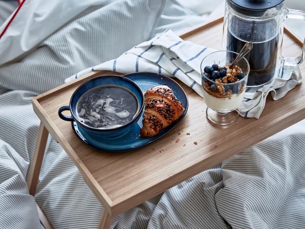 Sängyssä RESGODS-vuodetarjotin, jolle katettu kahvia, croissant ja jogurttia sekä marjoja jalkalasiin.