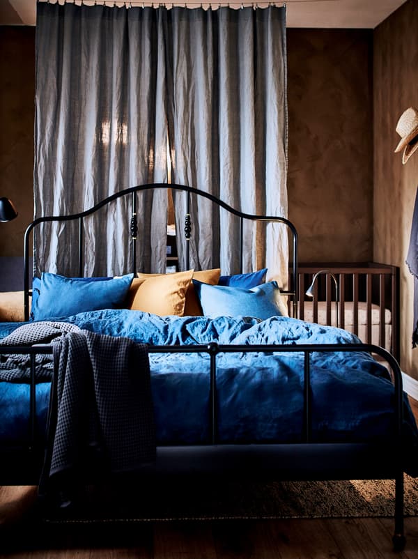 ห้องนอนที่มีเตียง SAGSTUA/ซ้ากสตัว สีดำและตรงมุมหลังผ้าม่านที่ใช้กั้นห้องมีเตียงเด็กอ่อนทำด้วยไม้ตั้งอยู่