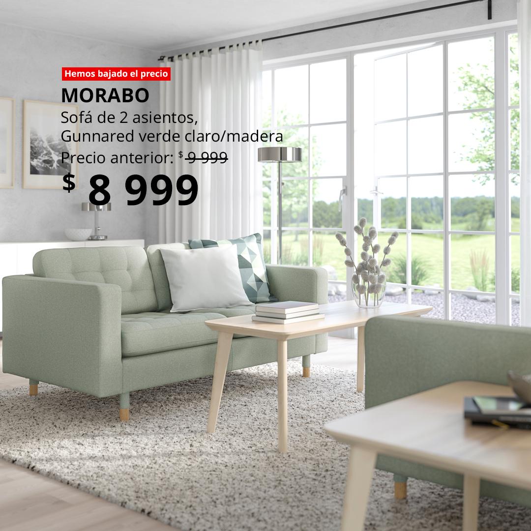 Sala contemporánea con sofá MORABO de 2 asientos verde claro Gunnared, patas de madera, mesa de centro de madera con libros y jarrón decorativo, alfombra gris y ventanales amplios con luz natural; diseño elegante, cómodo y minimalista.